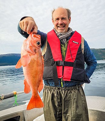 Ein Angler in Hitra, Norwegen, präsentiert stolz seinen Fang: einen orange-roten Fisch. Im Hintergrund malerische Fjordlandschaft bei bewölktem Himmel. Perfekt für Angelurlaub und unvergessliche Meeresangelerlebnisse.
