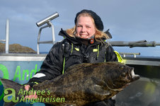 Eine Frau in wetterfester Kleidung hält einen großen Heilbutt auf einem Boot, im Hintergrund ist küstennahe Landschaft zu sehen. Eine Frau in wetterfester Kleidung hält einen großen Heilbutt auf einem Boot, im Hintergrund ist küstennahe Landschaft zu sehen.