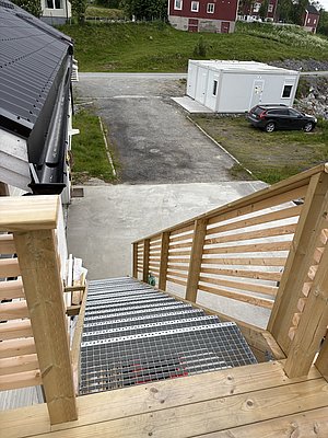 Blick von einer Holztreppe in Nord-Norwegen zeigt grünes Gras, eine asphaltierte Fläche, ein geparktes Auto und ein weißes Gebäude. Ideal für Angelreisende, die die raue Schönheit und Ruhe der Region genießen möchten.