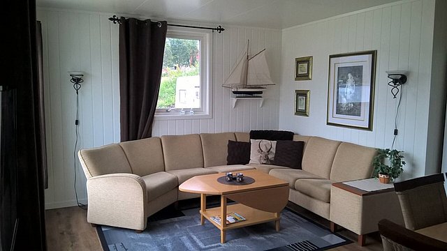 Gemütliches Wohnzimmer in Nord-Norwegen mit heller Eckcouch, stilvollen Wanddekorationen und maritimen Akzenten. Ideal für Entspannung nach dem Angelurlaub, bietet weiche Beleuchtung und einen einladenden Ausblick auf grüne Landschaft.