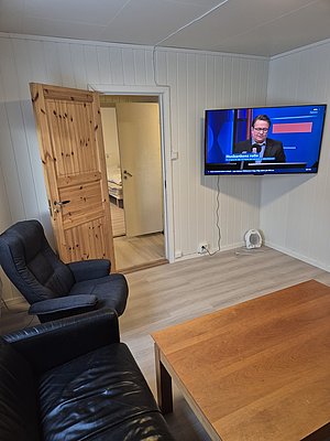 Ein gemütliches Ferienhaus in Nord-Norwegen mit modernen Annehmlichkeiten: Ledersessel, Holzregal, Fernseher an weißer Wand. Ideal für entspannende Angelreisen.
