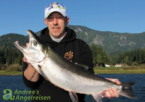 Ein Angler hält einen großen Lachs vor einer malerischen Berg- und Waldlandschaft in die Kamera.