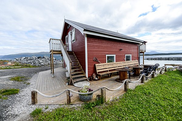 Rotes Ferienhaus in Nord-Norwegen direkt am Wasser mit Terrasse und Sitzbereich. Perfekt für einen erholsamen Angelurlaub mit malerischem Blick auf Berge und das Meer. Ideal für Natur- und Angelbegeisterte.