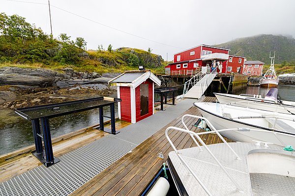 Rote Fischerhütten und Boote an einem ruhigen Steg im idyllischen Nordland, Norwegen. Perfekter Ausgangspunkt für Angelfreunde. Malerische Landschaft im typisch skandinavischen Stil, umgeben von grünen Hügeln und ruhigem Wasser.