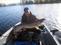 Ein Angler sitzt in einem Boot auf einem ruhigen See und hält einen großen Hecht stolz in die Kamera.