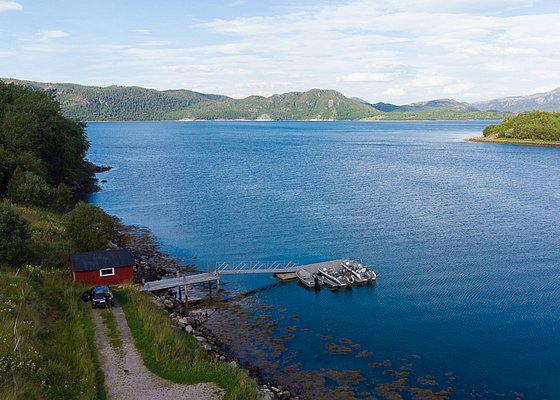 Malerische Küstenlandschaft Hitra, Norwegen: Blaues Meer, grüner Wald, kleiner Holzsteg mit Booten, rotes Häuschen. Perfekter Ausgangspunkt für Angelabenteuer und Naturerholung. Ideal für Entdecker und Angler auf Angelreisen in beeindruckender Naturkulisse.