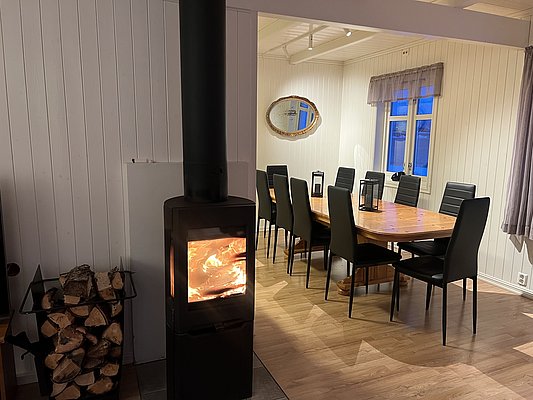 Gemütlicher Wohnraum in Nord-Norwegen mit Kaminofen, Holzvorrat, großem Esstisch mit schwarzen Stühlen und heller Holzwandverkleidung. Perfekte Atmosphäre für gesellige Angelabende nach einem erfolgreichen Tag am Meer.