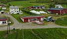 Luftaufnahme einer ländlichen Landschaft in Nord-Norwegen mit mehreren roten und weißen Gebäuden, grünen Wiesen, einer Schotterstraße und parkenden Fahrzeugen. Ideal für einen erholsamen Angelurlaub in idyllischer Naturumgebung.
