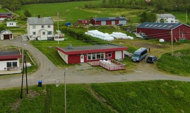 Luftaufnahme einer ländlichen Landschaft in Nord-Norwegen mit mehreren roten und weißen Gebäuden, grünen Wiesen, einer Schotterstraße und parkenden Fahrzeugen. Ideal für einen erholsamen Angelurlaub in idyllischer Naturumgebung.