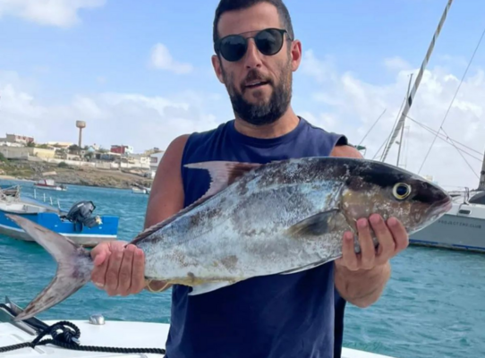 Ein Angler hält einen großen Fisch auf einem Boot vor der Küste von São Vicente, Kapverden. Im Hintergrund sind blaues Wasser und weitere Boote zu sehen – perfekter Ort für einen aufregenden Angelurlaub.