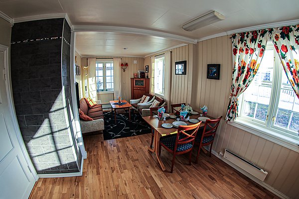 Gemütliches Wohnzimmer einer skandinavischen Ferienwohnung in Nord-Norwegen. Helle Holzböden, bunte Vorhänge, ein Esstisch und ein Sofa schaffen eine einladende Atmosphäre. Perfekt für Angelurlaub in malerischer Umgebung.