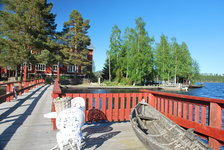 Holzsteg mit roten Geländern und weißen Gartenmöbeln führt zu einem traditionellen roten Gebäude am Seeufer, umgeben von Bäumen.