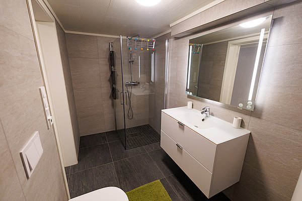 Modernes Badezimmer in Nord-Norwegen mit eleganter Dusche, großem Spiegel und stilvollem Waschtisch. Helle Fliesen und dezente Beleuchtung schaffen eine freundliche Atmosphäre. Ideal für einen komfortablen Aufenthalt in dieser reizvollen Region.