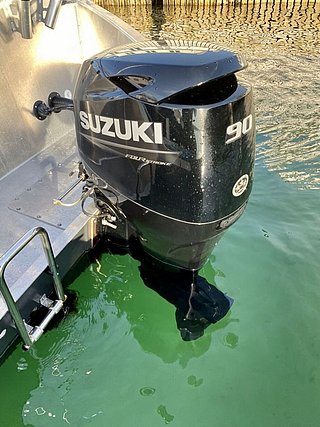 Ein Suzuki 90 PS Viertakt-Außenbordmotor am Heck eines Bootes über grünem Wasser.