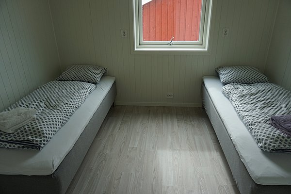 Gemütliches Schlafzimmer in Nordland, Norwegen: Zwei Einzelbetten mit gepunkteter Bettwäsche, helles Holzinterieur. Ein Fenster bietet Blick auf ein rotes Holzgebäude. Ideal für erholsamen Angelurlaub in skandinavischer Natur.