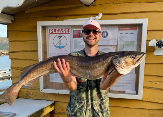 Ein Angler hält stolz einen großen Fisch vor einer gelben Holzhütte in Hitra, Norwegen. Im Hintergrund sind Informationsschilder und ein malerisches Küstenpanorama zu sehen. Perfektes Ziel für Angelreisen.