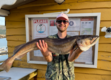 Ein Angler hält stolz einen großen Fisch vor einer gelben Holzhütte in Hitra, Norwegen. Im Hintergrund sind Informationsschilder und ein malerisches Küstenpanorama zu sehen. Perfektes Ziel für Angelreisen.
