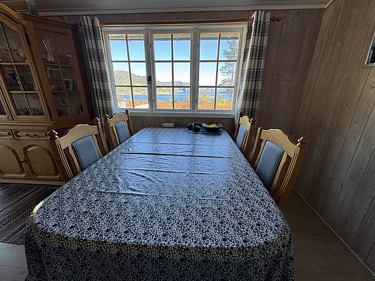 Gemütliches Esszimmer in Hitra, Norwegen, mit Holzmöbeln. Blick aus dem Fenster auf malerische Landschaft und fjordartige Küste. Ideal für Angelreisende, die komfortables Wohnen mit atemberaubenden Ausblicken kombinieren möchten.