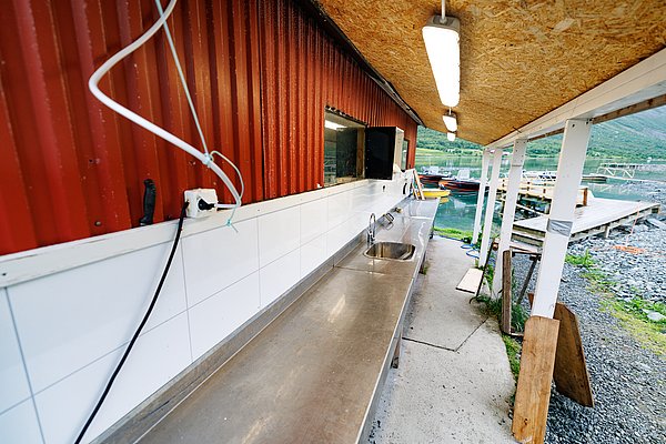 Außenbereich eines Fischverarbeitungsstandes in Nord-Norwegen, mit Edelstahl-Arbeitsfläche und Spüle, neben einem malerischen Hafen mit Booten – idealer Startpunkt für Angelreisen.