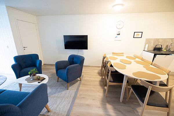 Gemütliches Wohnzimmer in Nordland, Norwegen: Blaue Sessel, moderner Fernseher, großer Esstisch und Küchenzeile. Ideal für Angelurlaub mit entspanntem Ambiente. Perfekter Ort zum Erholen nach einem erfolgreichen Tag am Wasser.