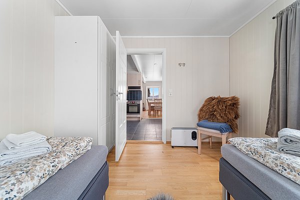 Gemütliches Schlafzimmer in Nord-Norwegen mit zwei Einzelbetten, Holzmöbeln und sanfter Beleuchtung. Der offene Durchgang zeigt das einladende Wohnzimmer mit Blick auf den Fjord. Ideal für Angelurlaub und Erholung in skandinavischer Natur.