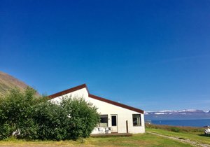 Weißes Haus in Sudavik, Island, vor beeindruckender Fjordlandschaft. Blauer Himmel, umliegende grüne Natur und ruhige Wasserkulisse bieten reizvolle Angelabenteuer. Ideal für Angelreisen und Urlaube in unberührter, malerischer Umgebung.