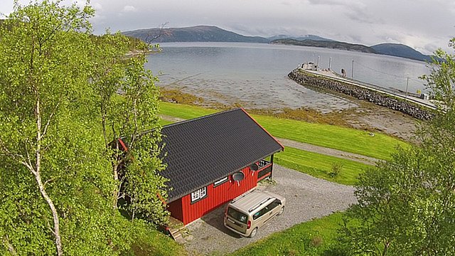 Rotes Blockhaus am Fjord in Nordland, Norwegen. Umgeben von grüner Natur, Blick auf das ruhige Wasser und Berge im Hintergrund. Perfekt für Angelurlaub, Entspannung und Naturerlebnis. Ideal für Erholungssuchende und Abenteurer.