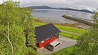 Rotes Blockhaus am Fjord in Nordland, Norwegen. Umgeben von grüner Natur, Blick auf das ruhige Wasser und Berge im Hintergrund. Perfekt für Angelurlaub, Entspannung und Naturerlebnis. Ideal für Erholungssuchende und Abenteurer.
