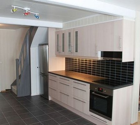 Moderne Küche in Nord-Norwegen mit hellen Holzschränken, schwarzer Arbeitsplatte und Kacheln. Edelstahl-Kühlschrank, Herd und bunte Deckenleuchten. Ideal für komfortable Aufenthalte und Angelreisen in Skandinavien.