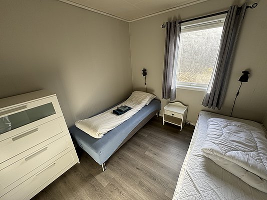 Gemütliches Schlafzimmer in Hitra, Norwegen: Zwei Betten, Fenster mit Vorhängen, weiße Kommode und Nachttisch. Ideal für einen erholsamen Angelurlaub mit idyllischem Blick auf die Natur. Perfekte Basis für Abenteuer und Entspannung.