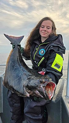 Anglerin hält beeindruckenden Heilbutt auf einem Boot in Nord-Norwegen. Perfektes Ziel für Angelreisen mit starken Fangchancen und atemberaubender Naturkulisse. Ideal für alle, die Abenteuer und Entspannung suchen.