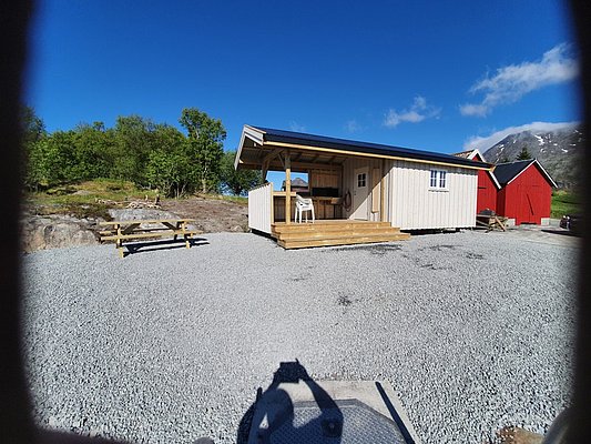 Gemütliche Holzhütte in Nordland, Norwegen, umgeben von grünen Bäumen und blauem Himmel. Perfekt für Angelurlaube. Barrierefrei zugänglich mit Terrasse und Picknicktischen. Ideal für Naturliebhaber und ruhige Erholung.