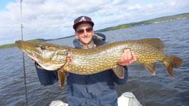 Eine Person hält einen großen Hecht vor einem See, während sie auf einem Boot steht, unter einem bewölkten Himmel.