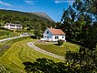 Gemütliches weißes Haus in Nord-Norwegen mit rotem Dach, umgeben von grüner Landschaft und Bergen. Perfekter Ausgangspunkt für Angelreisen und Naturabenteuer. Strahlend blauer Himmel ergänzt die idyllische Szenerie.