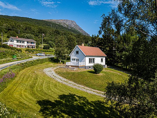Gemütliches weißes Haus in Nord-Norwegen mit rotem Dach, umgeben von grüner Landschaft und Bergen. Perfekter Ausgangspunkt für Angelreisen und Naturabenteuer. Strahlend blauer Himmel ergänzt die idyllische Szenerie.
