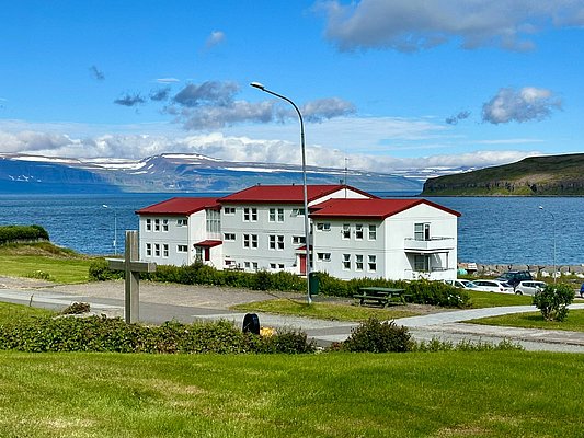 Weißes Gebäude mit rotem Dach in Sudavik, Island. Malerische Landschaft mit Bergen und Meer im Hintergrund, grüner Rasen im Vordergrund. Perfekter Ort für Erholung und Naturliebhaber. Ideal für Angelreisen und Entdeckungstouren.