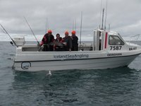 Eine Gruppe von Menschen steht auf einem Boot namens "Odínshani" mit der Aufschrift "Iceland Sea Angling" und hält einen großen Fisch, umgeben von Angelruten und Möwen über einem wolkigen Ozean.