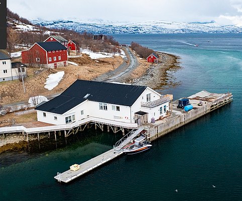 Küstenlandschaft in Nord-Norwegen mit weißen und roten Häusern am Fjord, umgeben von schneebedeckten Bergen. Ein idealer Ausgangspunkt für Angelabenteuer in beeindruckender Naturkulisse. Perfekt für Angelreisen und Erholung.