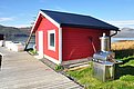 Rotes Holzhaus in Nord-Norwegen am Wasser, umgeben von beeindruckender Berglandschaft. Ein Steg führt direkt zur Tür und ein Grill steht bereit. Perfekt für Angler und Naturliebhaber. Ideal für Angelurlaube und Erholung.