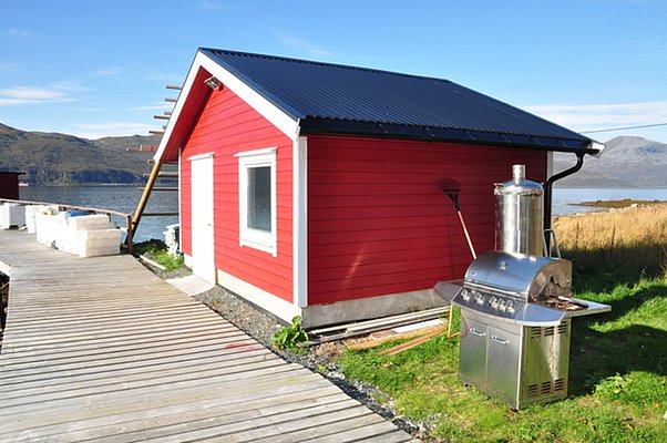 Rotes Holzhaus in Nord-Norwegen am Wasser, umgeben von beeindruckender Berglandschaft. Ein Steg führt direkt zur Tür und ein Grill steht bereit. Perfekt für Angler und Naturliebhaber. Ideal für Angelurlaube und Erholung.