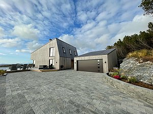 Modernes Ferienhaus in Hitra, Norwegen, mit weitläufigem Pflastersteinhof, umgeben von malerischer Natur und Meerblick unter bewölktem Himmel. Ideal für einen erholsamen Angelurlaub in skandinavischer Idylle.