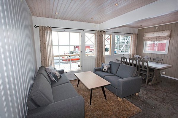 Gemütliches Wohnzimmer in nordnorwegischer Ferienhütte, mit grauen Sofas, Holztisch und Esstisch. Helle Fenster mit Blick auf den Hafen. Perfekt für entspannte Angelurlaub-Tage in Nord-Norwegen. Ideal für Erholung nach einem erfolgreichen Angeltag.
