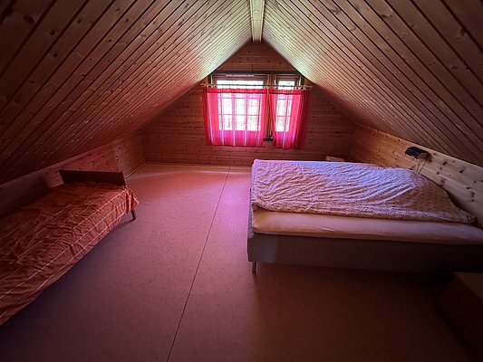 Gemütliches Dachzimmer in Hitra, Norwegen, mit Holzwänden, kleinem Fenster mit roten Vorhängen, Doppel- und Einzelbett. Ideal für ruhige Angelurlaub-Aufenthalte in naturnaher Umgebung. Perfekt zum Entspannen nach erfolgreichen Angeltouren.