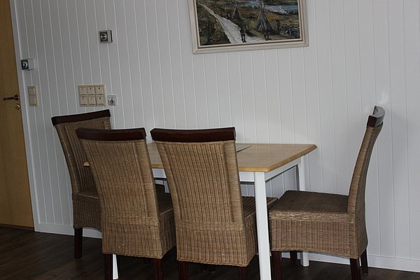 Essbereich einer skandinavischen Unterkunft in Nord-Norwegen mit Holztisch, vier geflochtenen Stühlen und heller Wandverkleidung. Nordisches Gemälde an der Wand verleiht dem Raum eine gemütliche Atmosphäre. Ideal für Angelreisen in Norwegen.