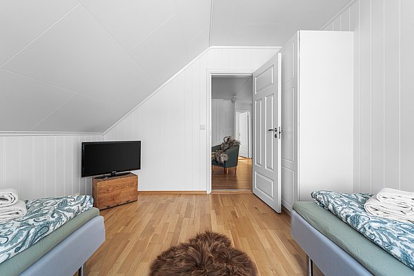 Helles Schlafzimmer in Nord-Norwegen mit zwei Betten, Holzboden und TV. Offene Tür führt zu Wohnbereich mit Sessel. Gemütliche Unterkunft für Angelreisende, ideal zum Entspannen nach einem erfolgreichen Angeltag.