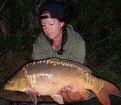 Eine Person hält einen großen Karpfen in den Händen und posiert bei Nacht vor einer mit Büschen bewachsenen Kulisse.