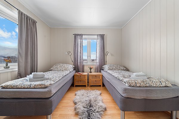 Gemütliches Schlafzimmer mit zwei Einzelbetten, hellen Holzwänden und Fenstern mit Bergblick in Nord-Norwegen. Ideal für entspannten Angelurlaub. Flauschiger Teppich auf Holzboden verleiht Wärme und Komfort.
