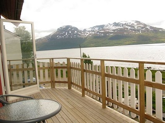 Holzterrasse mit Tisch und Stühlen, Blick auf norwegische Fjordlandschaft und schneebedeckte Berge. Perfekter Ort für Entspannung und Naturgenuss in Nord-Norwegen. Ideal für Angelreisen und Erholung.