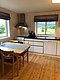 Gemütliche, helle Küche in Nord-Norwegen mit Holzfußboden, Esstisch, Stühlen und moderner Ausstattung. Große Fenster bieten Blick auf grüne Landschaft und Berge. Perfekt für Angelurlaub in skandinavischer Ruhe.