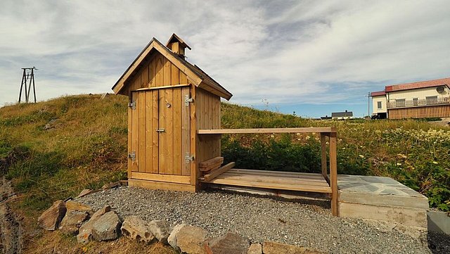 Kleines, hölzernes Gebäude auf Kiesboden in der idyllischen Landschaft Nordlands, Norwegen. Der blaue Himmel ergänzt die ruhige, naturnahe Umgebung. Perfekt für Angelreisende, die unberührte Natur und Ruhe suchen.